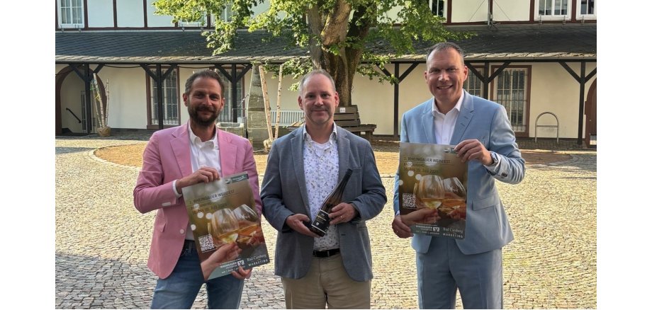 Zu sehen ist: Dominik Russler, Kurdirektor Michael Sinn und Bürgermeister Daniel Rühl im Amthof-Innenhof mit dem Weinfest-Plakat